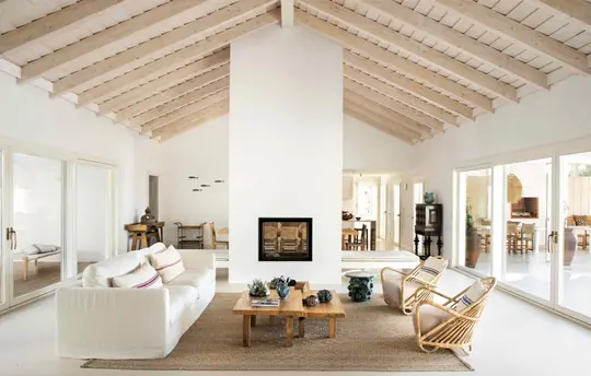 Salón reformado con vigas de madera vistas, chimenea blanca y sofá de lino sobre alfombra de yute